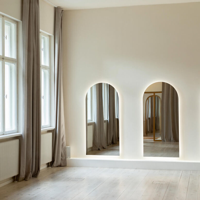A minimalistic room with double arched mirrors on the wall and soft light streaming in through the windows.