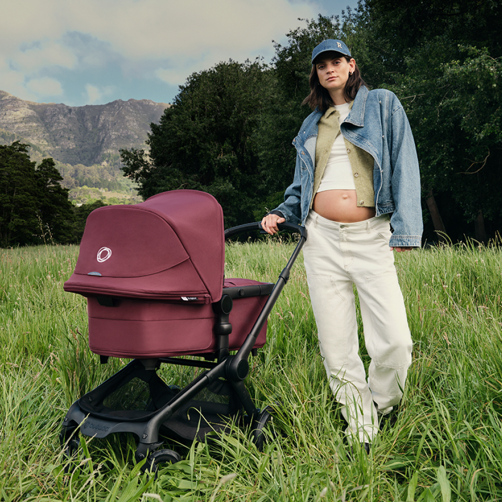 Een moeder in casual streetwear met een zichtbare zwangere buik staat in een grasveld met een Bugaboo Fox 5 Renew kinderwagen, met bomen en bergen op de achtergrond.