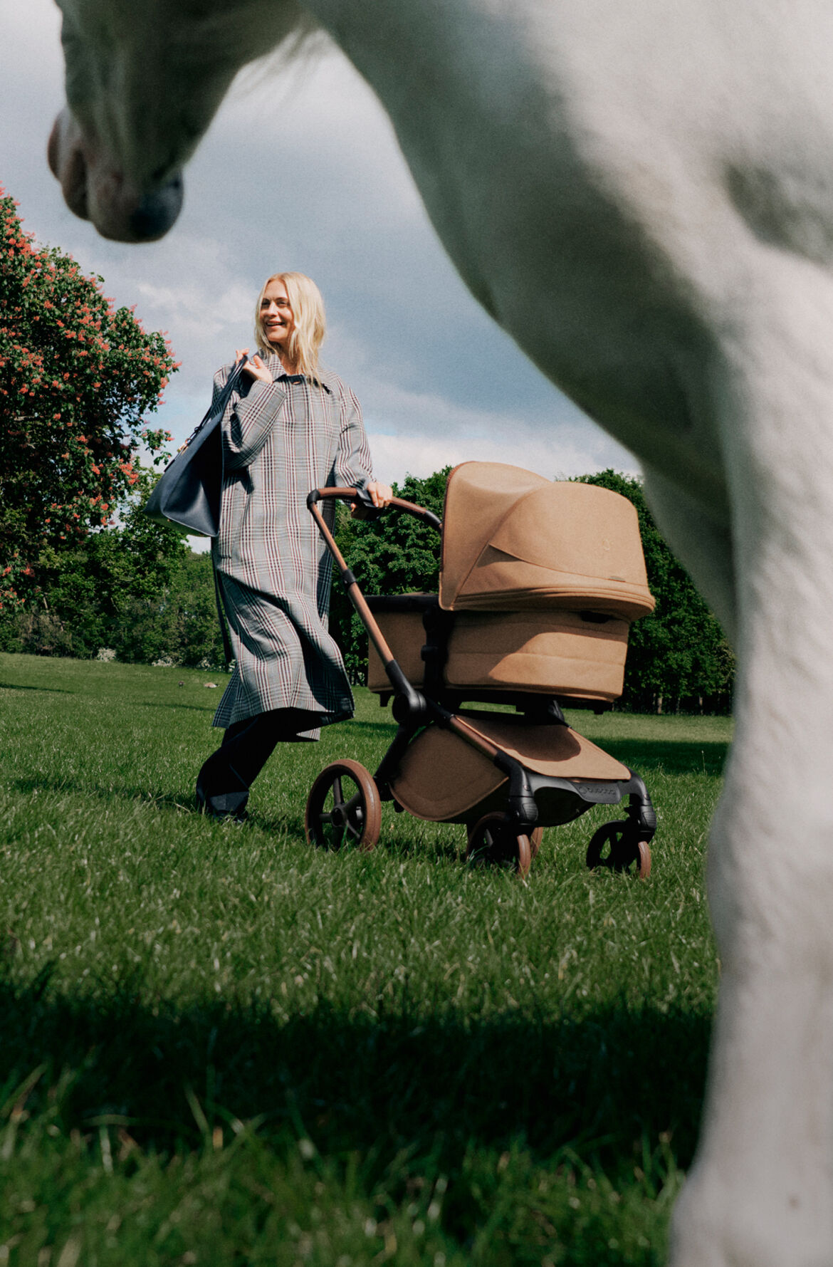 Une maman élégante pousse une poussette Bugaboo x Stella McCartney dans un parc avec des arbres verdoyants et un ciel bleu en arrière-plan. Au premier plan, partiellement hors cadre et flou, se trouve un poney blanc.