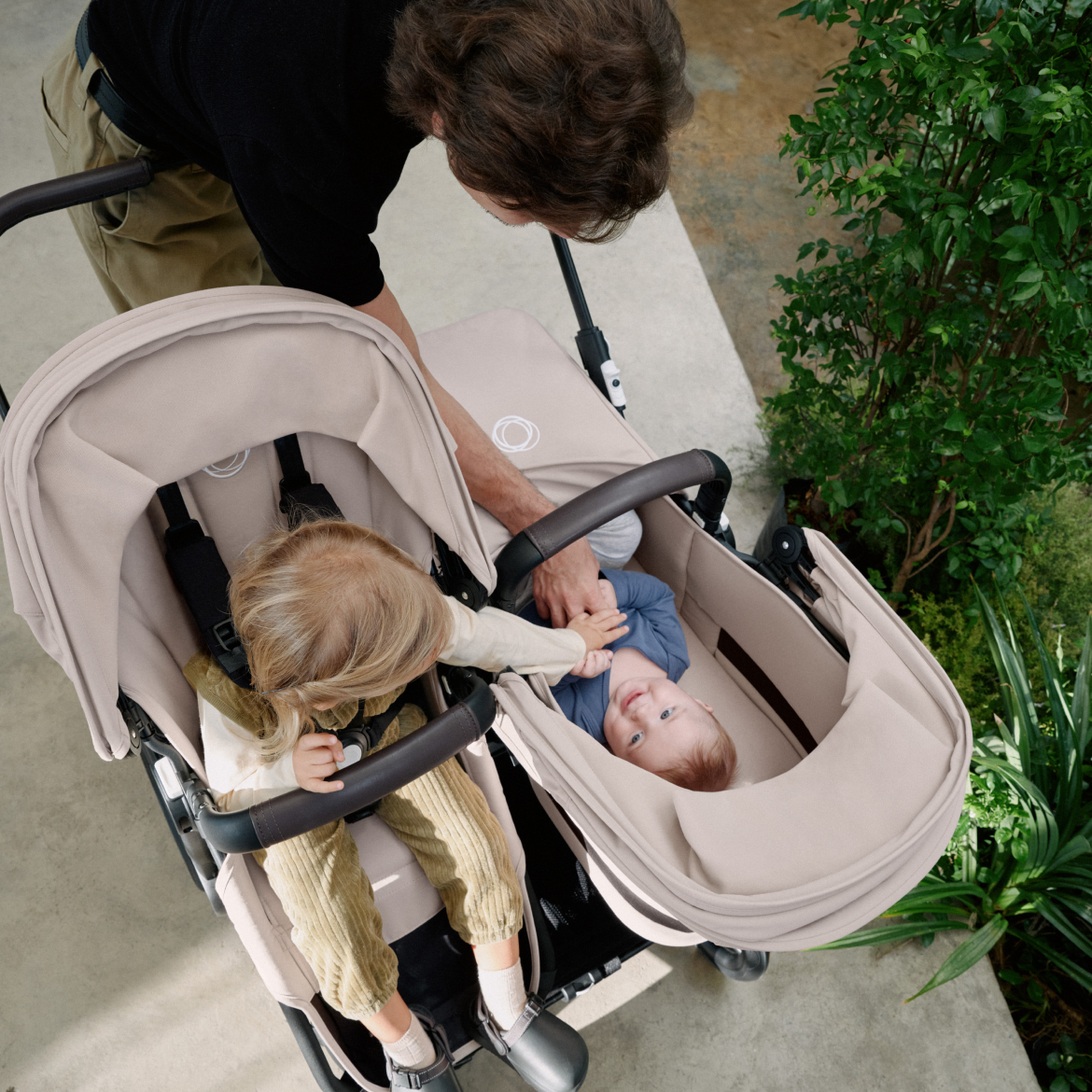 A mom strolls with two children in a Bugaboo Donkey 5 double pram. The girl in the seat is reaching out to her sibling in the bassinet.