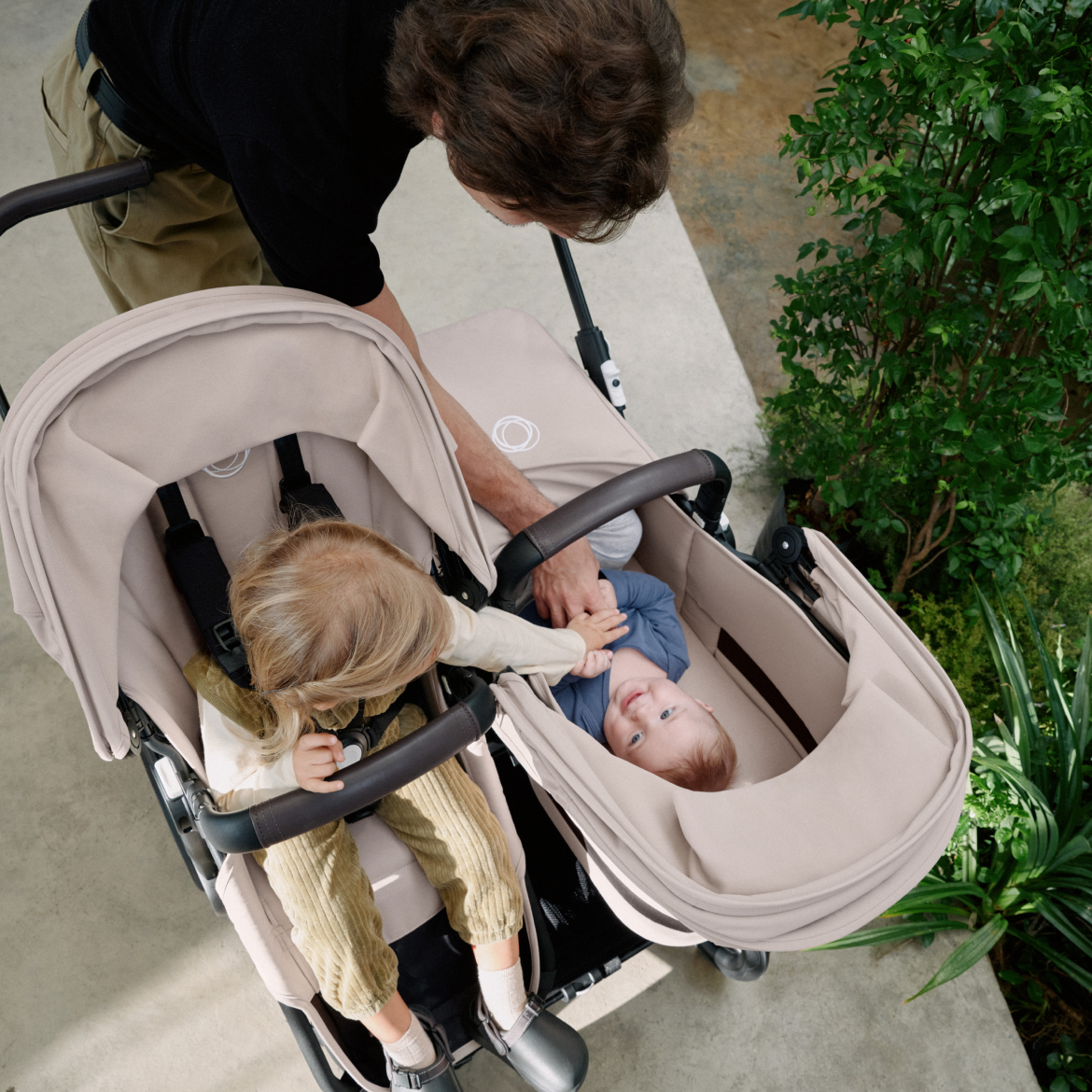 Een moeder wandelt met twee kinderen in een Bugaboo Donkey 5 duo kinderwagen. Het meisje in de stoel strekt zich uit naar de baby in de wieg.