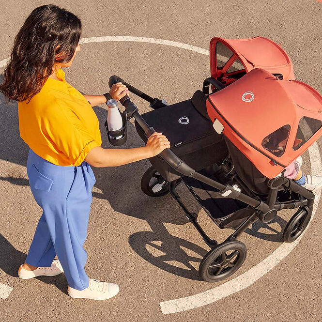 Mom pushing a Bugaboo double pushchair, with a bottle in a cup holder attached to the pushchair's handlebar.