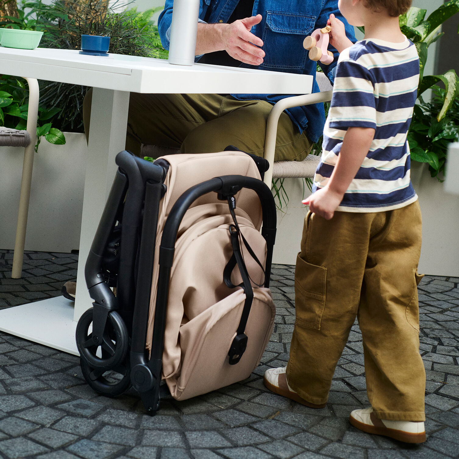 A Bugaboo Butterfly 2 Plus travel stroller folded compactly and stands upright next to a restaurant table. A toddler stands next to it.