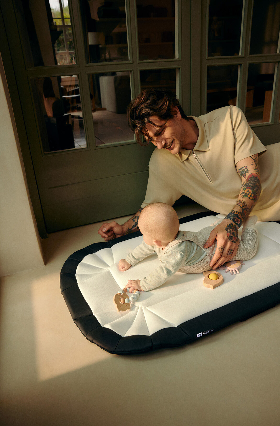 A dad gently supports his baby as she plays on a padded Bugaboo baby nest used as a playmat. They are at home, sitting together on the floor of a hallway.