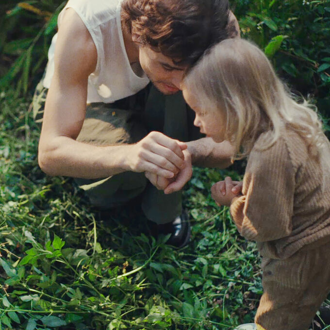 Een vader en dochter hurken in een ongetemde, overwoekerde tuin. Hij laat haar iets zien dat hij in zijn handen heeft.