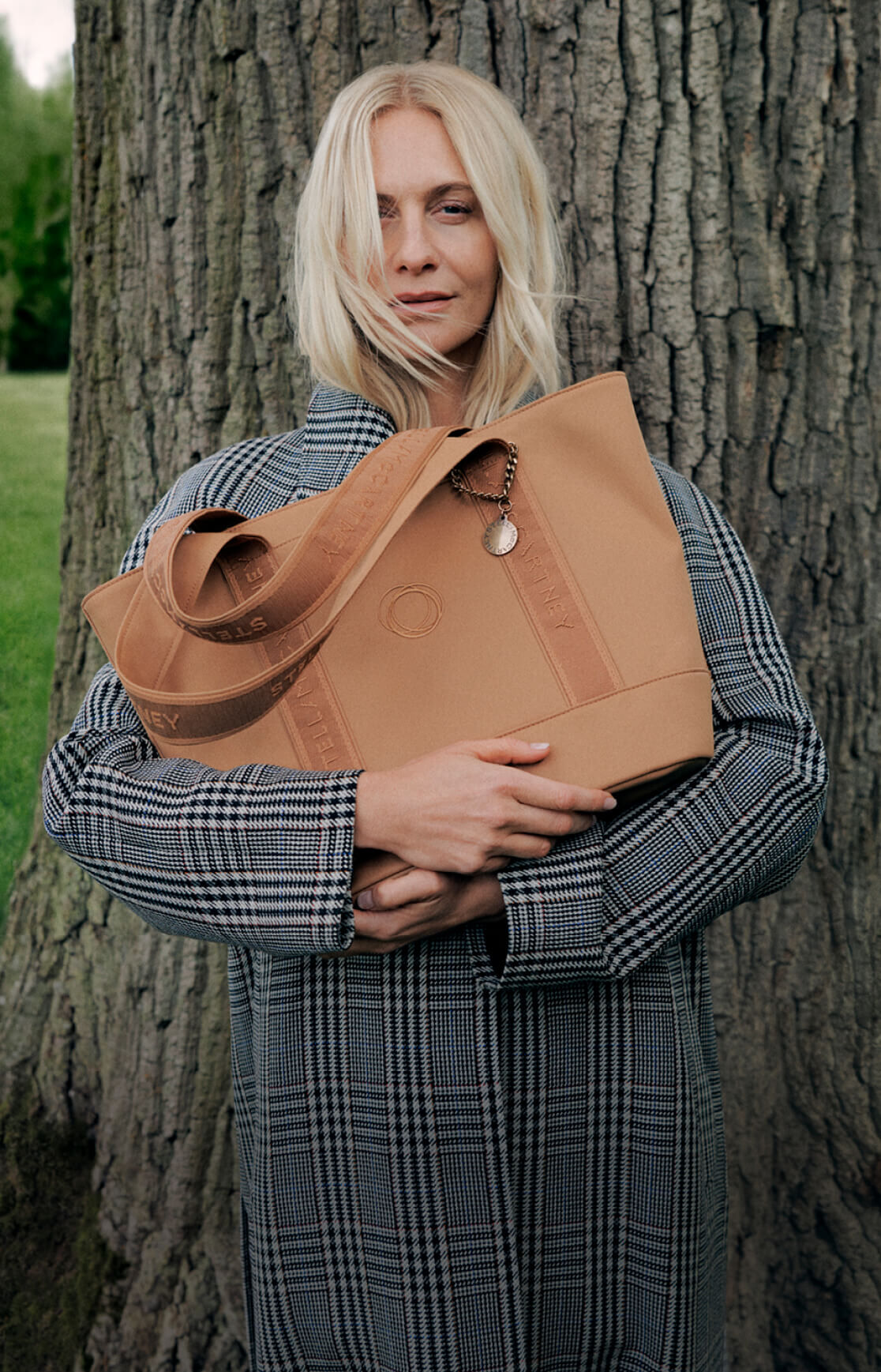 Poppy Delevingne leans against a large tree trunk while holding a Bugaboo x Stella McCartney tote bag.