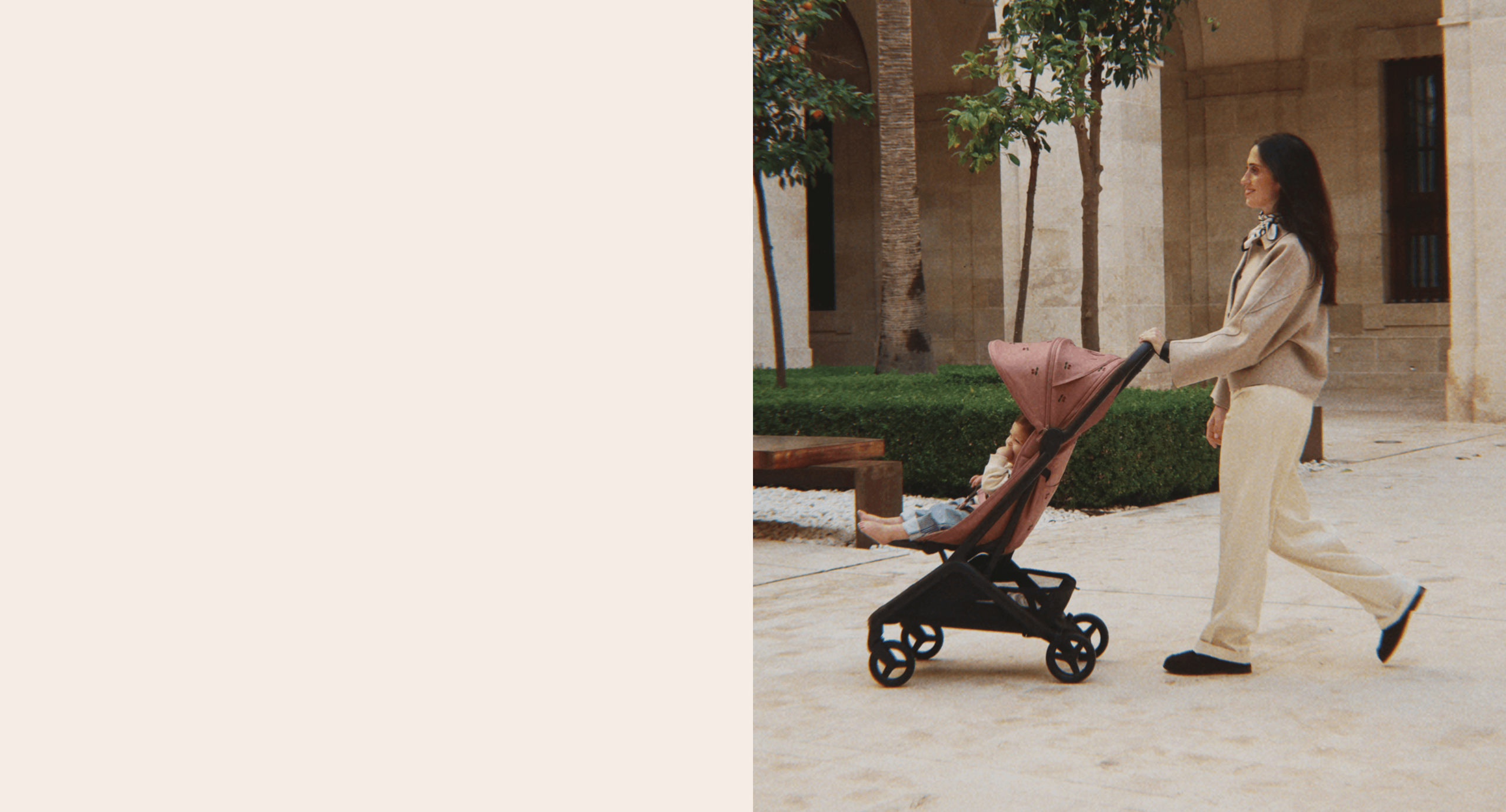 A smiling mom walks with her baby sitting in a Bugaboo Butterfly 2 stroller. The stroller features limited-edition fabric embroidered with Konges Sløjd cherry motif. They’re in a green courtyard outside a stone-walled building.