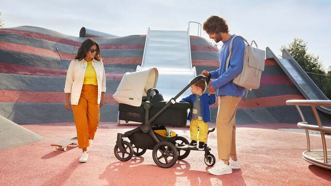 A family of four enjoying a day out; the father is carrying a Bugaboo changing backpack and pushing a Bugaboo stroller, while a toddler is standing on a Bugaboo wheeled board