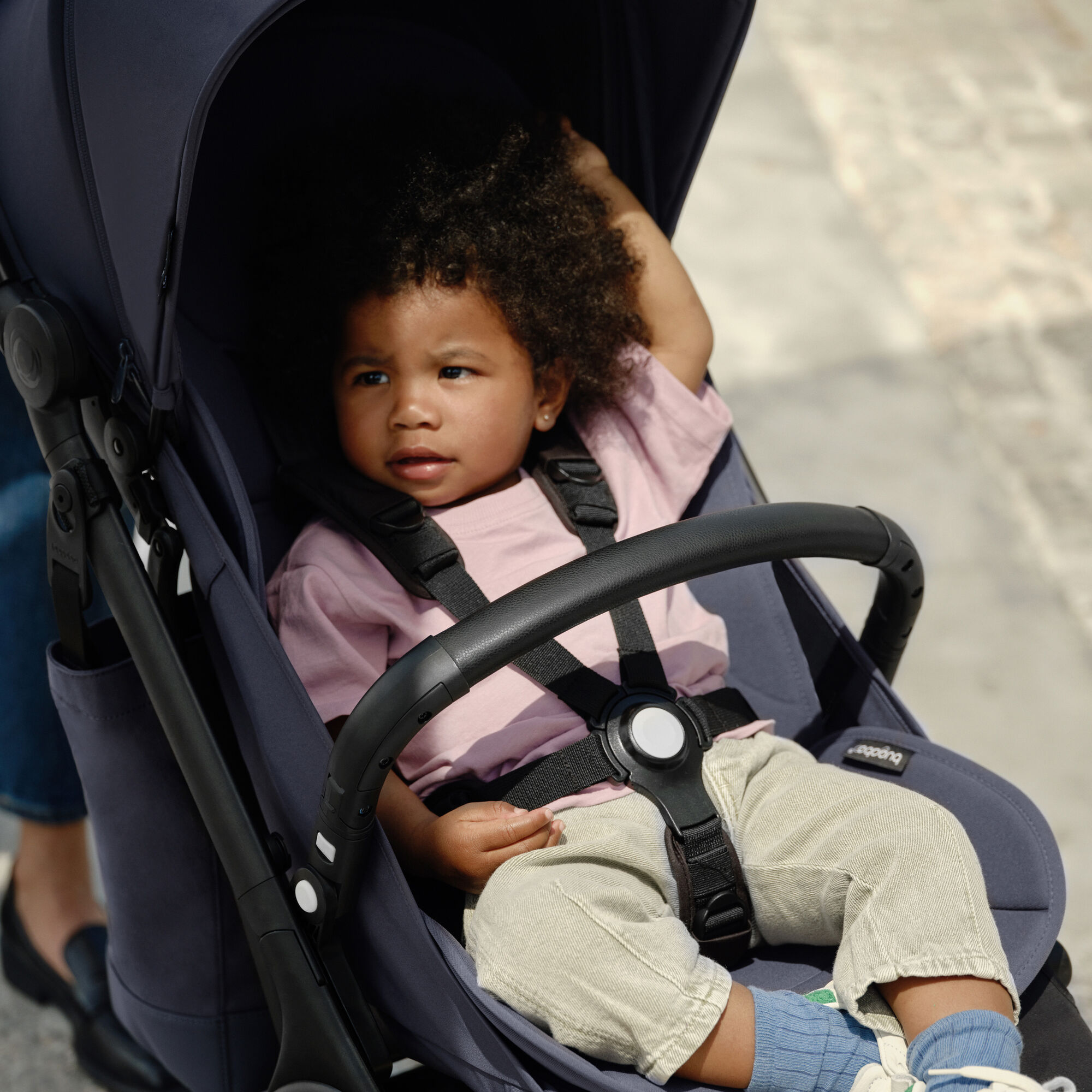 A toddler relaxes in the seat of an ultra-compact Bugaboo Butterfly travel pram. The toddler is securely seated with the harness fastened and a Bugaboo Butterfly bumper bar across their middle. 