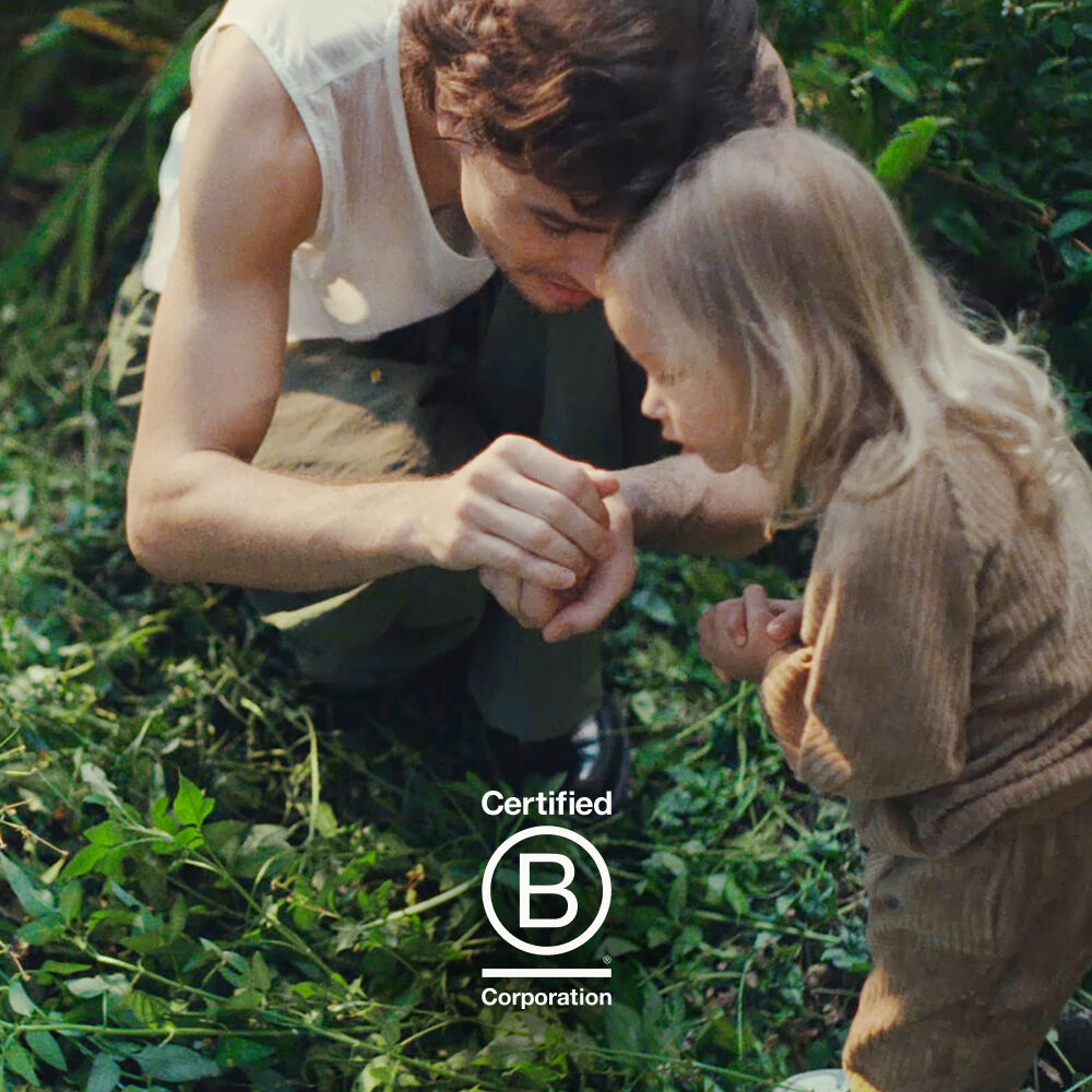 A dad and his daughter crouch in an untamed, overgrown garden. He's showing her something he's caught in his hands. The Certified B Corp logo sits at the bottom of the image.