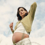 A pregnant mom stands in a grassy field with one hand on her bare baby bump, with mountains in the background and tall blades of grass in the foreground.