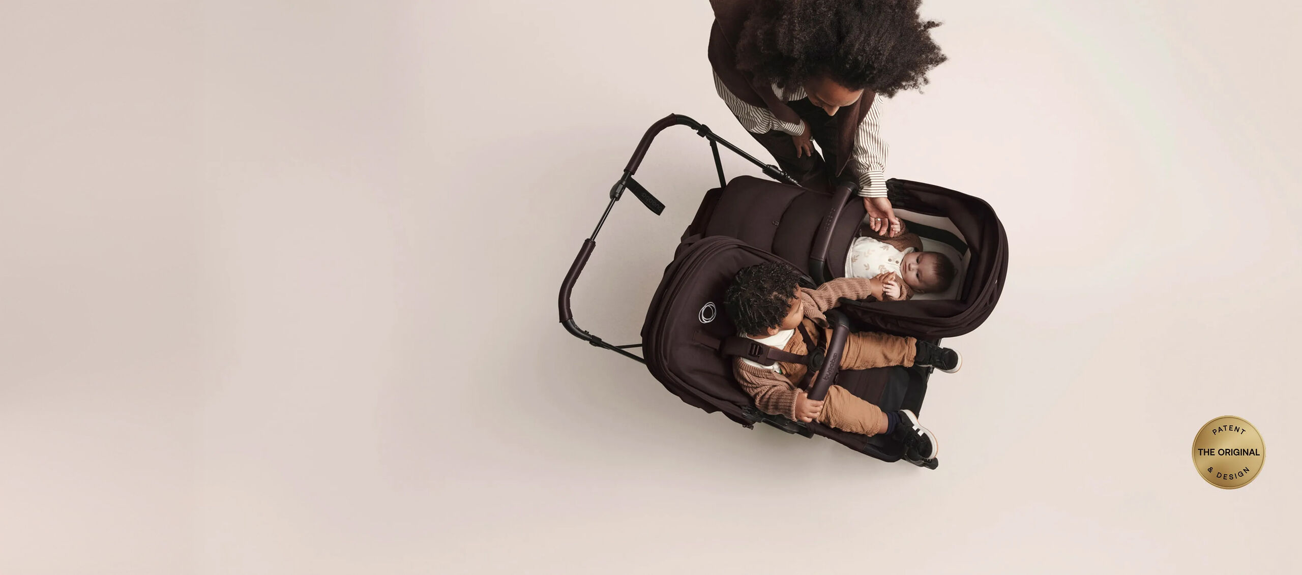 Blick von oben auf den Bugaboo Donkey 6 Kinderwagen in der Doppelkonfiguration. Das Kleinkind auf dem Sitz streckt die Hand nach seinem kleinen Geschwisterchen in der Liegewanne aus. Ihre Mama steht neben ihnen und streichelt das Baby sanft. Unten rechts ist ein gelber runder Stempel mit der Aufschrift „The Original Patent & Design“ (Das Originalpatent und -design).