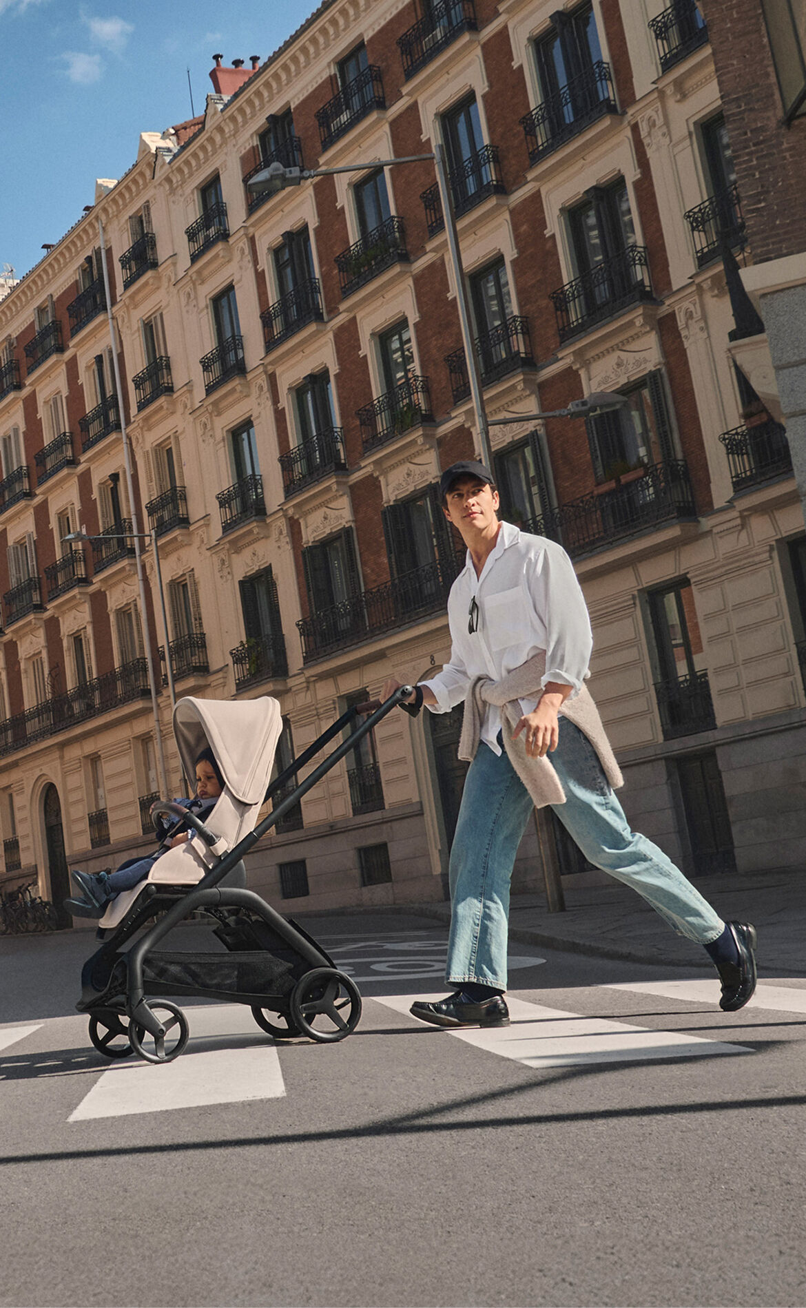 A dad pushes a Bugaboo Dragonfly Plus stroller with a child, crossing a city street lined with tall apartment buildings on a sunny day.