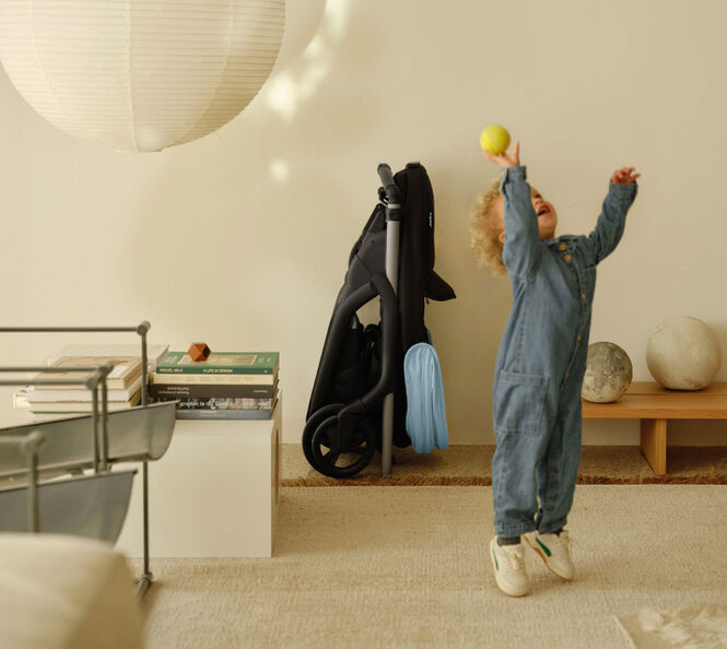 Ett litet barn leker i ett modernt vardagsrum och kastar en boll i luften. I bakgrunden syns en Bugaboo Dragonfly-barnvagn, med sufflett i blå färg (Skyline Blue). Barnvagnen är kompakt ihopfälld och står prydligt lutad mot en vägg.