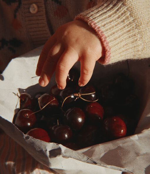 A tiny baby’s hand reaches into a bowl full of red cherries.