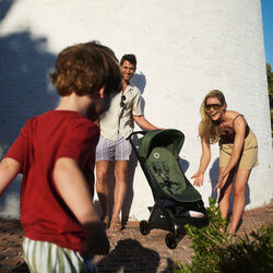 A baby sits barefoot in the sand with his mom behind him, on the beach near large rocks on a sunny day.