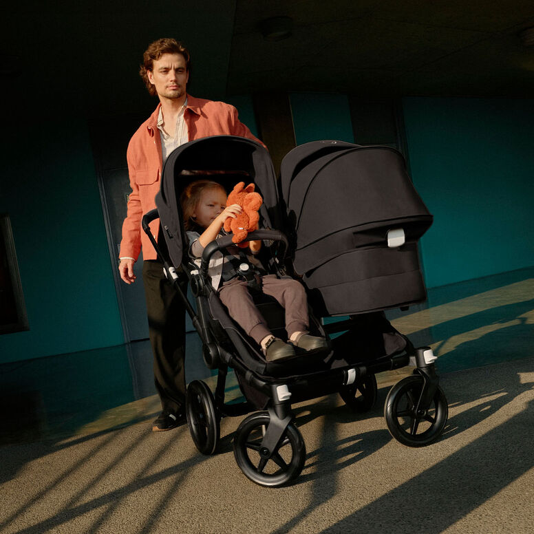 A dad makes a turn with a Bugaboo Donkey 5 Duo stroller. His toddler is in the seat facing the world and holding a stuffed animal. The bassinet is facing the dad