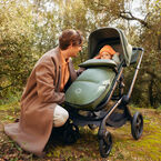A father kneels beside his baby in a Bugaboo stroller on an autumnal forest path. The baby is bundled up in a cozy Bugaboo THERMOLITE ® performance footmuff. They are surrounded by tall trees with soft sunlight filtering through the foliage.