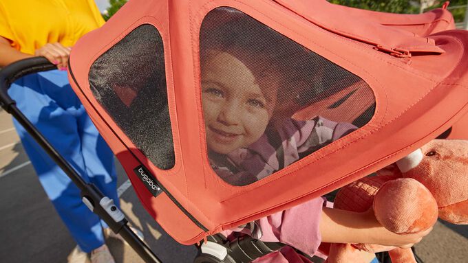 A toddler plays with her toy in a Bugaboo stroller while smiling at the camera from behind a red Bugaboo breezy sun canopy