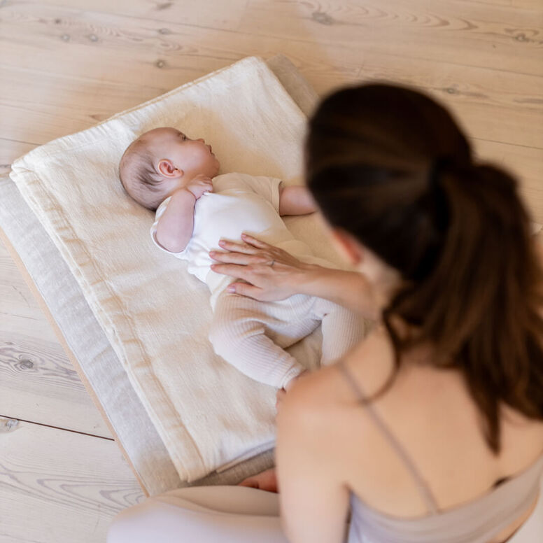 A mother sits on the floor, touching her baby's belly who is lying on a towel in front of her. They are seated in a brightly lit space.