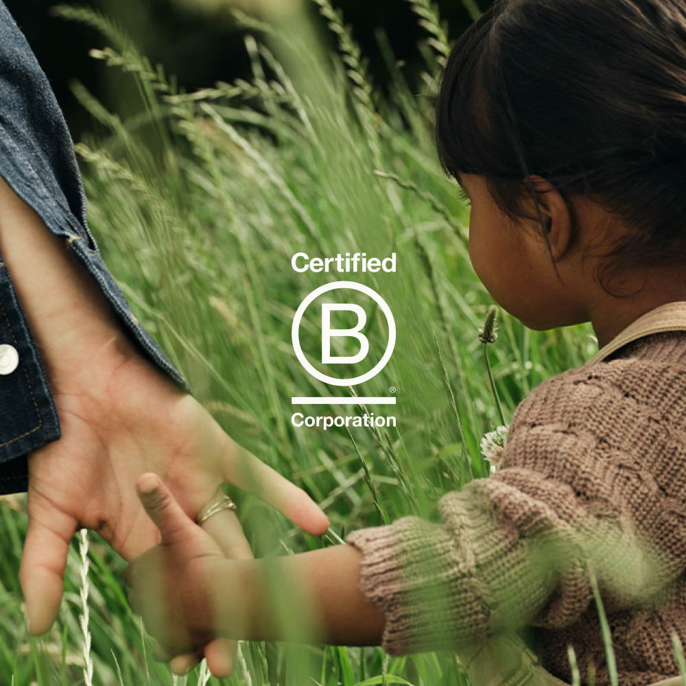 Een peuter loopt door het hoge gras terwijl ze de hand van haar moeder vasthoudt. Over de afbeelding is een logo geplaatst met de tekst 'Certified B Corporation'.