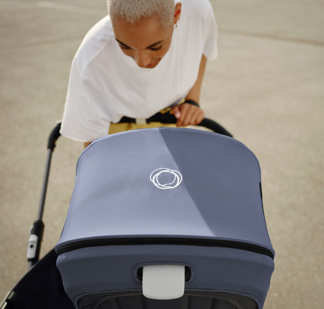 A mother leans over her Bugaboo pram to check on her baby. The Bugaboo pram has a Stormy Blue canopy, with a white Bugaboo logo in the centre.