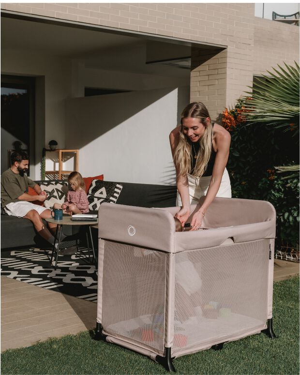 A mom leans down to pick up her baby from a Bugaboo Stardust travel cot, set in a garden and featuring Desert Taupe fabrics. In the background, a dad and daughter sit and play on a sofa.