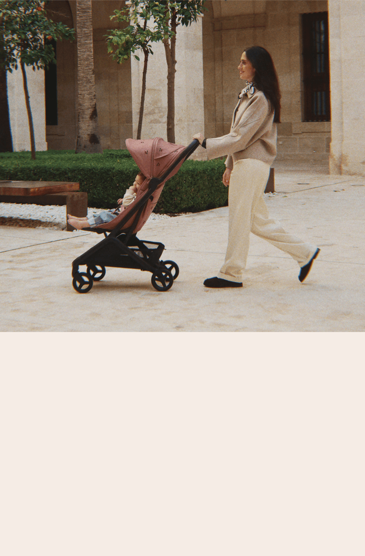 A smiling mom walks with her baby sitting in a Bugaboo Butterfly 2 stroller. The stroller features limited-edition fabric embroidered with Konges Sløjd cherry motif. They’re in a green courtyard outside a stone-walled building.