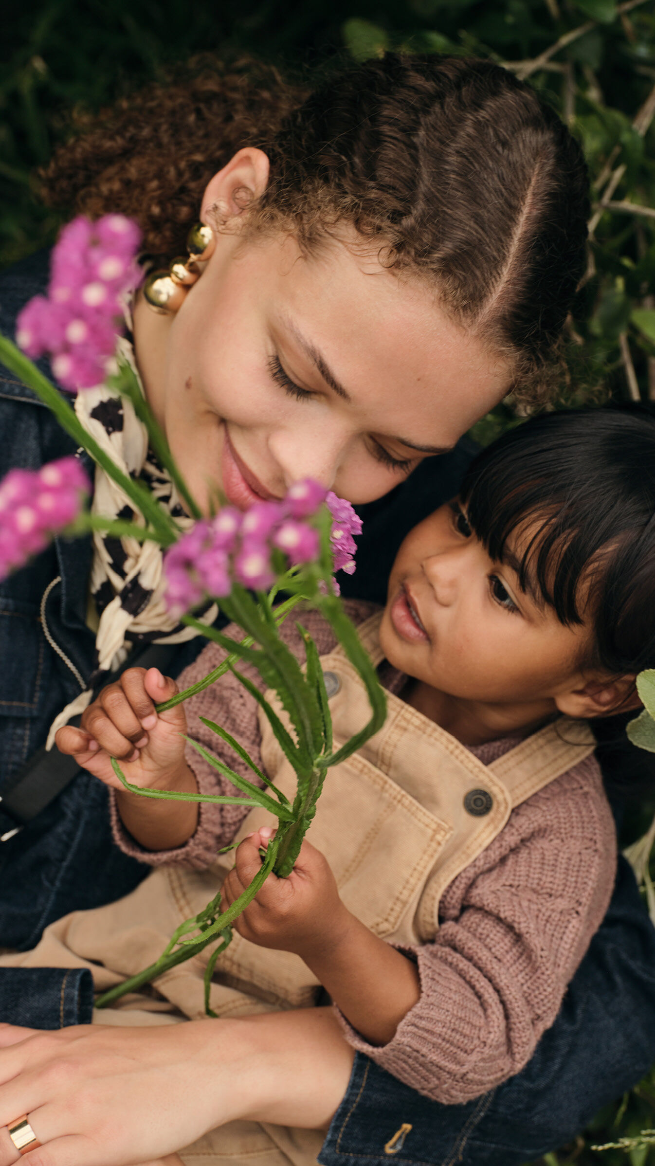 A smiling mom and her daughter sit close together, gently holding and examining a small bouquet of purple flowers.