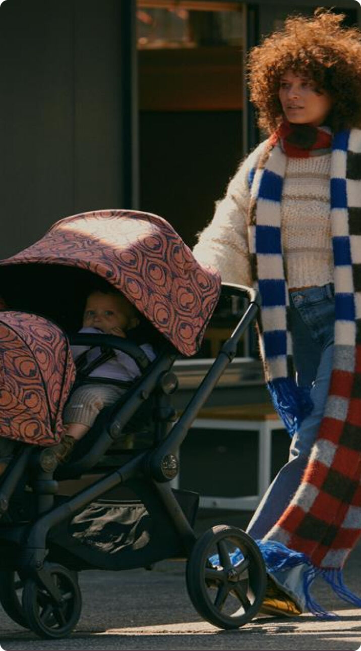 A mom wearing a checkered scarf strolls with her children seated in a Bugaboo Kangaroo tandem stroller. The stroller features Bugaboo X Artipoppe Limited Edition sun canopy.