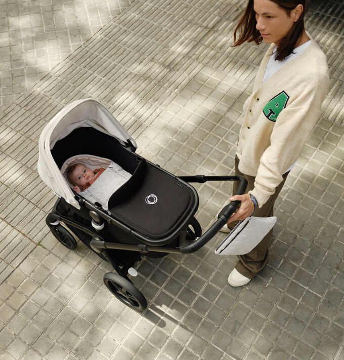 Un padre in piedi accanto al figlio neonato in un passeggino Bugaboo. Il passeggino è dotato di navicella, di un sacco navicella e di una pochette per il cambio Bugaboo.