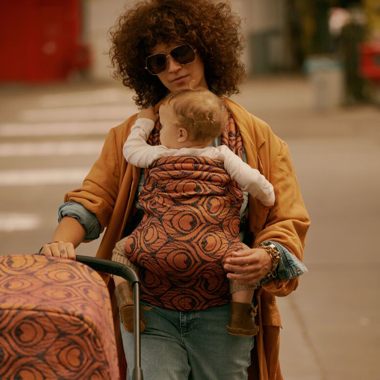 A stylish mom carries her baby in an Artipoppe Zeitgeist Baby Carrier. She’s holding onto a Bugaboo stroller with the Bugaboo X Artipoppe Limited Edition sun canopy. Behind them is a casual, urban setting.