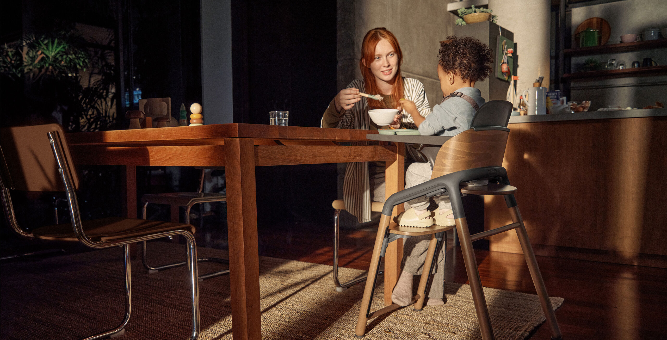 A mom feeds her toddler who’s seated in a Bugaboo Giraffe high chair with a tray attached. They’re sitting in a warm, softly lit kitchen with wooden furniture.