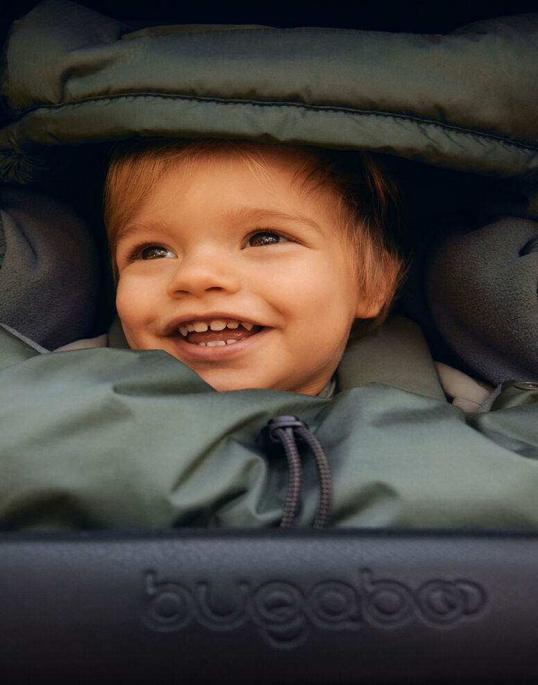 A baby smiling brightly as they're cuddled up in a footmuff inside a stroller.