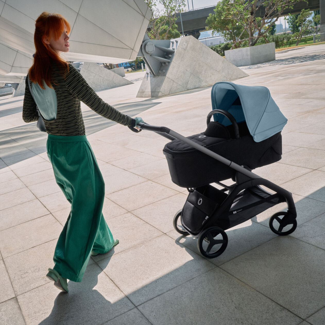 Une maman élégante se promène avec son bébé dans une Bugaboo Dragonfly en passant devant un bâtiment au loin duquel se trouve un jardin.