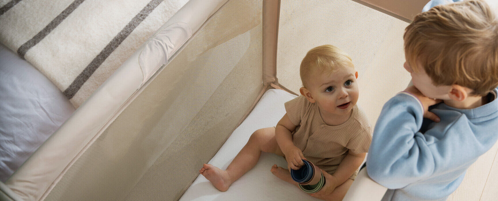 Top view of a baby napping peacefully inside a Bugaboo Stardust play yard. Next to the play yard, a toddler is playing at a low table.