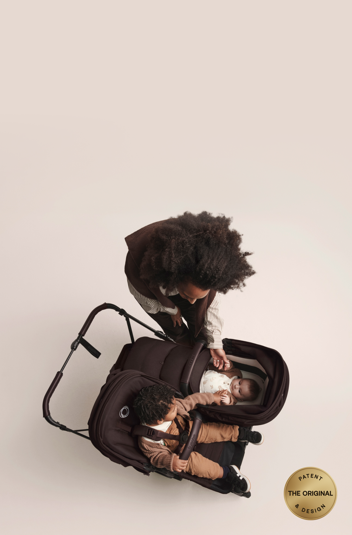 Blick von oben auf den Bugaboo Donkey 6 Kinderwagen in der Doppelkonfiguration. Das Kleinkind auf dem Sitz streckt die Hand nach seinem kleinen Geschwisterchen in der Liegewanne aus. Ihre Mama steht neben ihnen und streichelt das Baby sanft. Unten rechts ist ein gelber runder Stempel mit der Aufschrift „The Original Patent & Design“ (Das Originalpatent und -design).