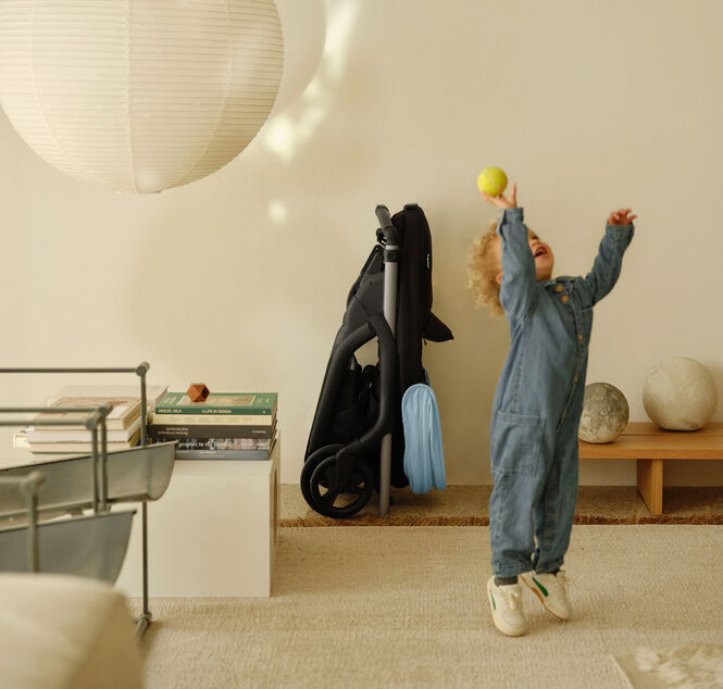 A toddler plays in a contemporary living room, throwing a ball into the air. In the background, a Bugaboo Dragonfly city pushchair, with a Skyline Blue canopy, is compactly folded and tucked away neatly against a wall.
