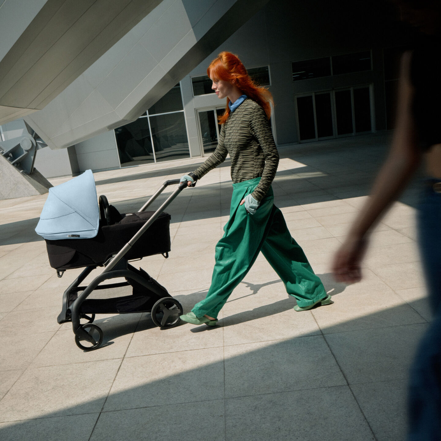 Een zelfverzekerde moeder loopt met haar baby in een Bugaboo Dragonfly stadswandelwagen terwijl ze langs een futuristisch gebouw glijdt
