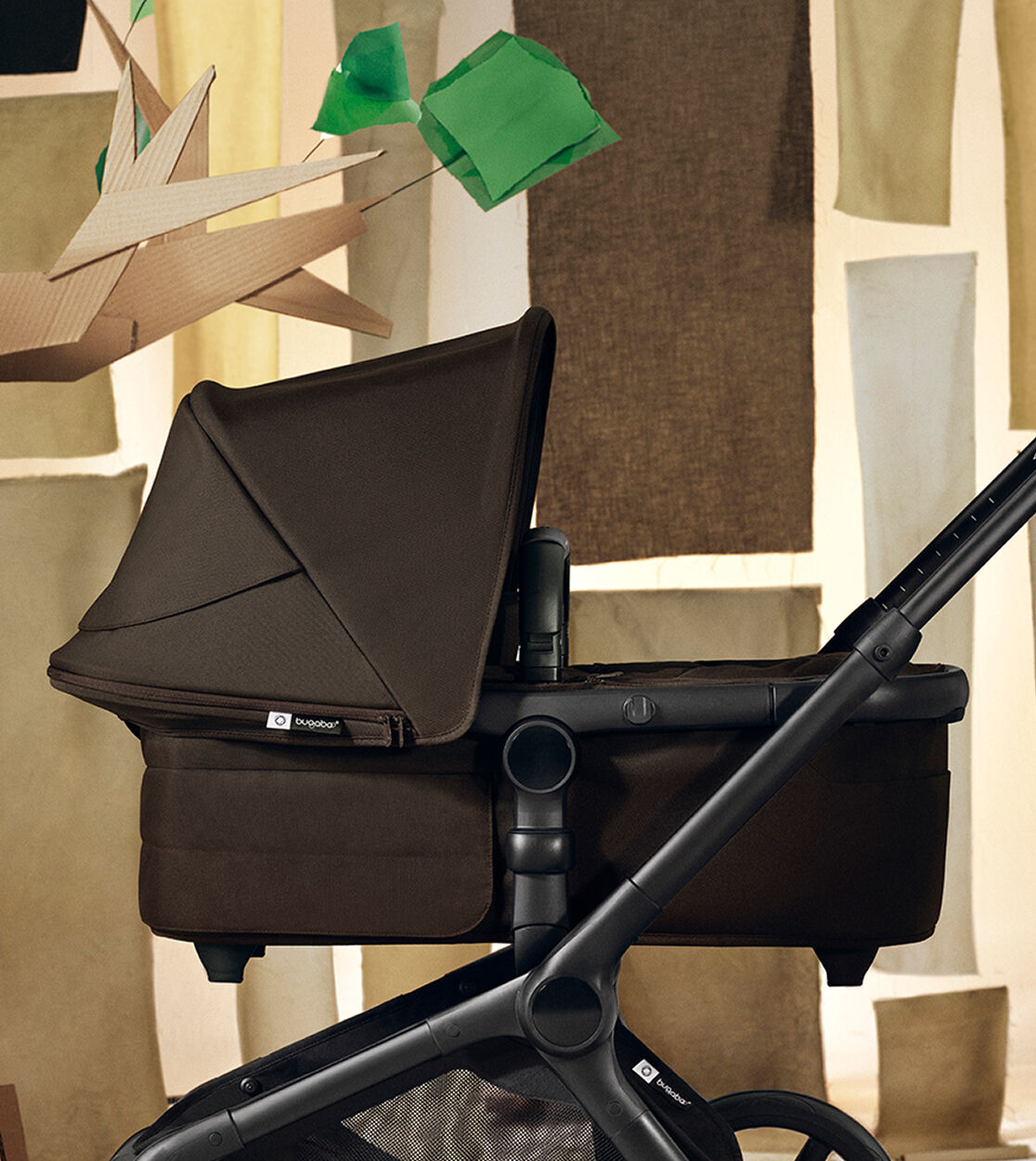 A Bugaboo Fox 5 Renew seat stroller in Fern Green and a Bugaboo Fox 5 Renew bassinet stroller in Cocoa Brown. They’re in a stylized indoor set with a cardboard tree and abstract city-building shapes.