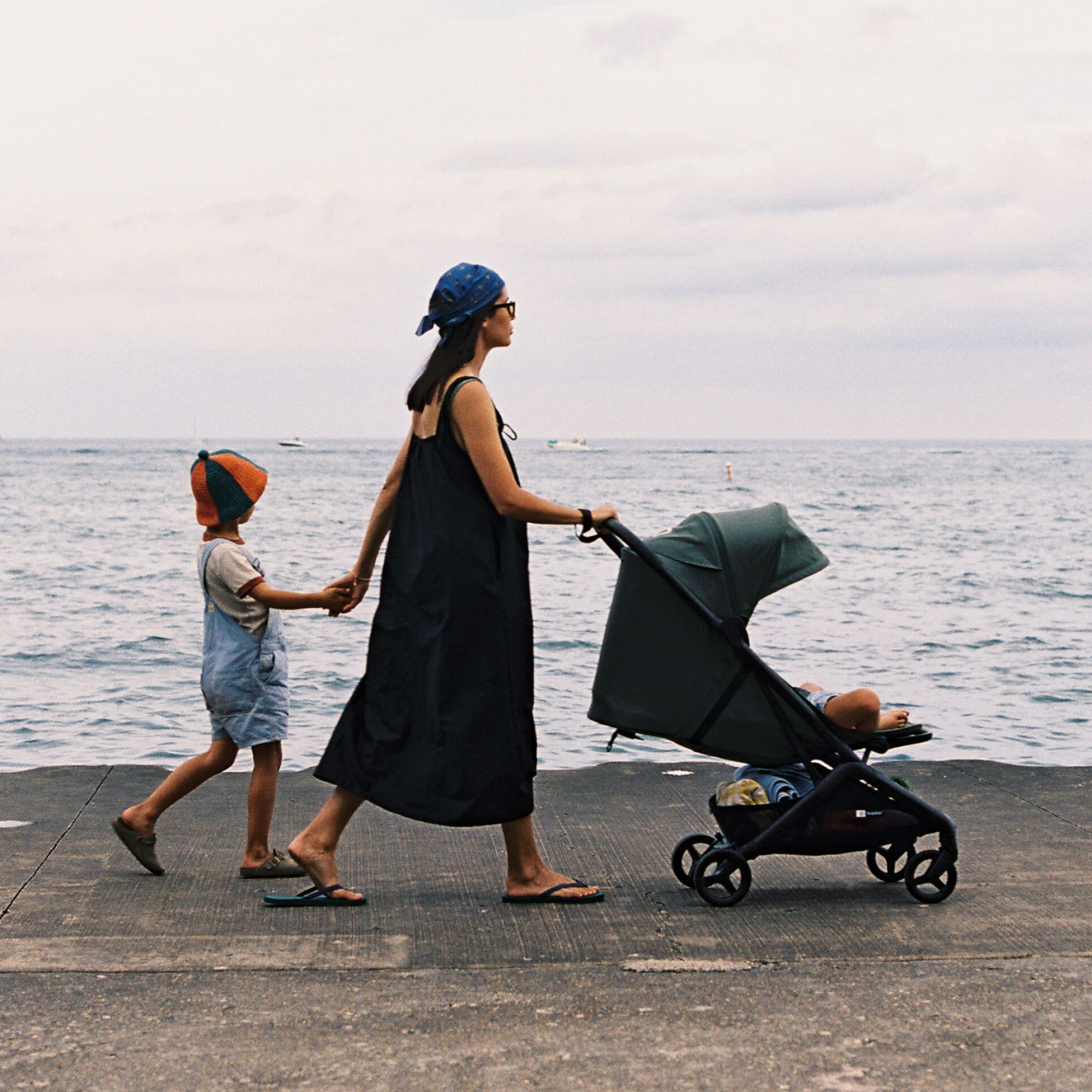 Mom and child walk hand in hand while mom pushes a Bugaboo Butterfly 2 Plus travel stroller with a toddler inside. They are strolling by the sea.