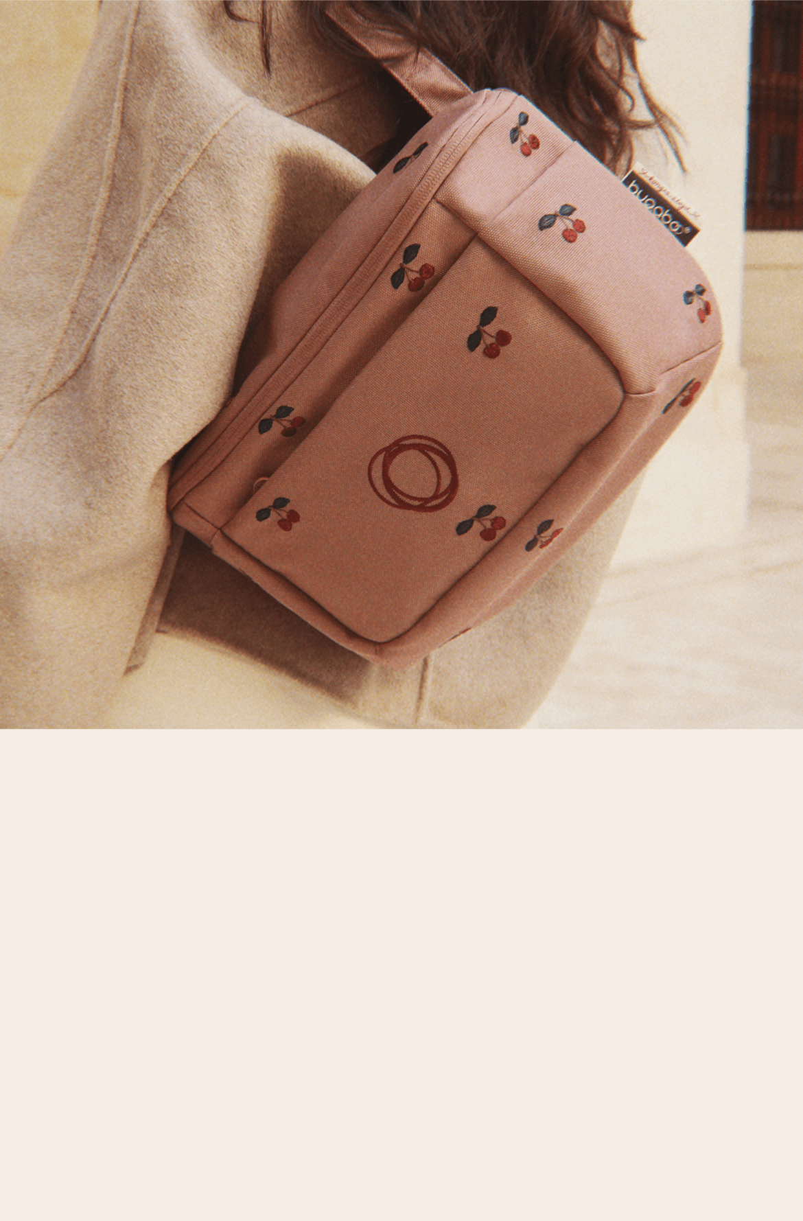 A close up of a Bugaboo x Konges Sløjd Limited Edition organizer, with cherry patterns embroidered on Dusty Pink fabric, worn over a beige coat.