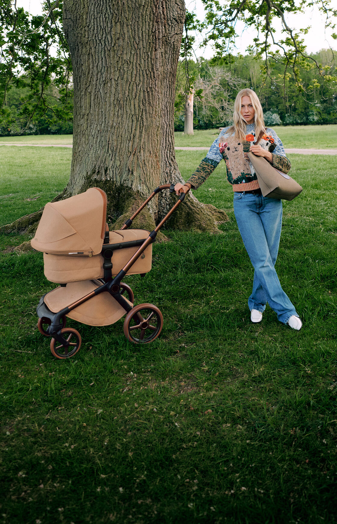 Poppy Delevingne stands next to a Bugaboo x Stella McCartney Fox 5 Renew pushchair, holding a bag and wearing a knitted jumper. Behind her is a large tree in a grassy field.
