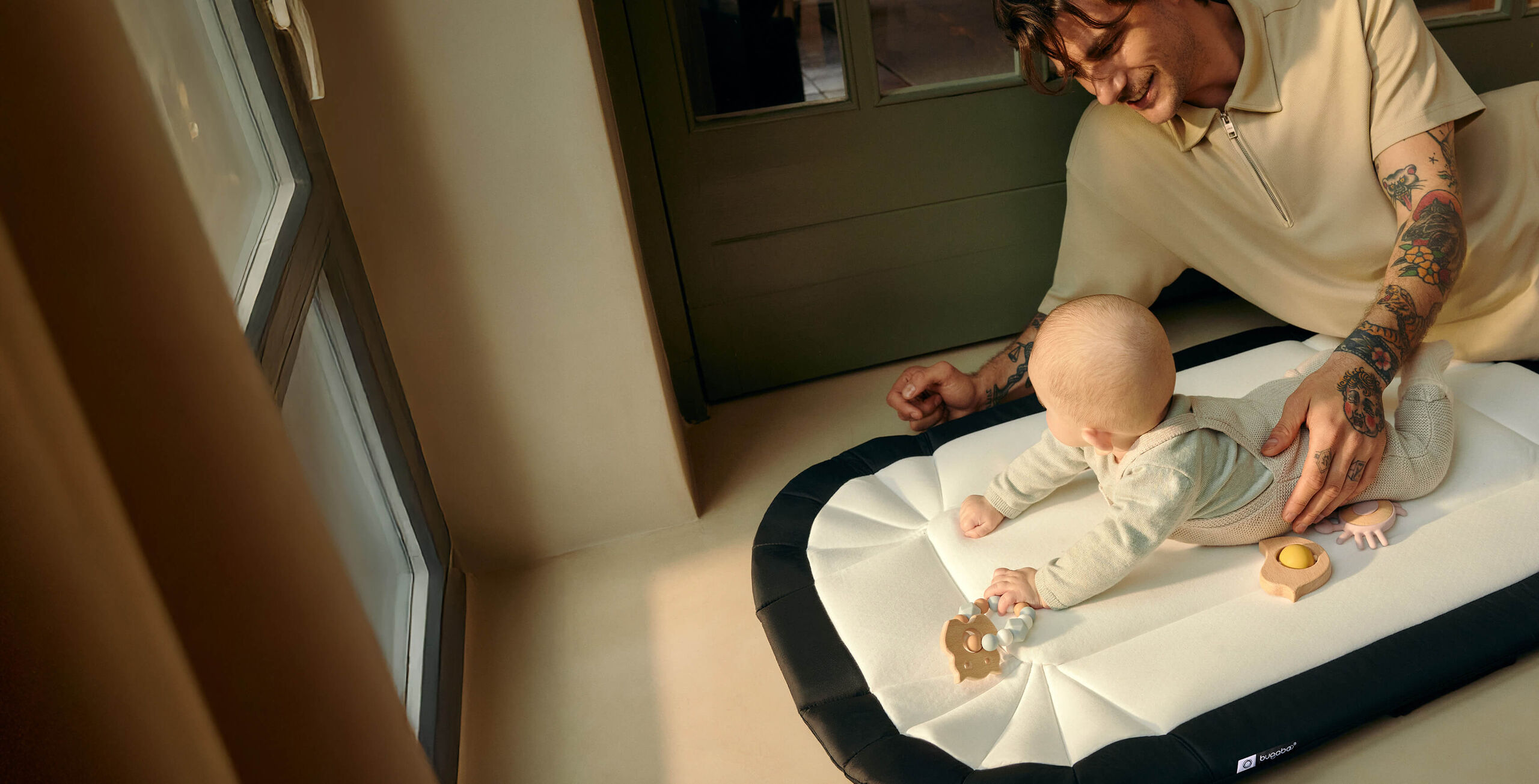 A dad gently supports his baby as she plays on a padded Bugaboo baby nest used as a playmat. They are at home, sitting together on the floor of a hallway.