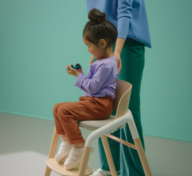 A child sitting on the Bugaboo Giraffe while playing with a toy, with her Mom standing behind.