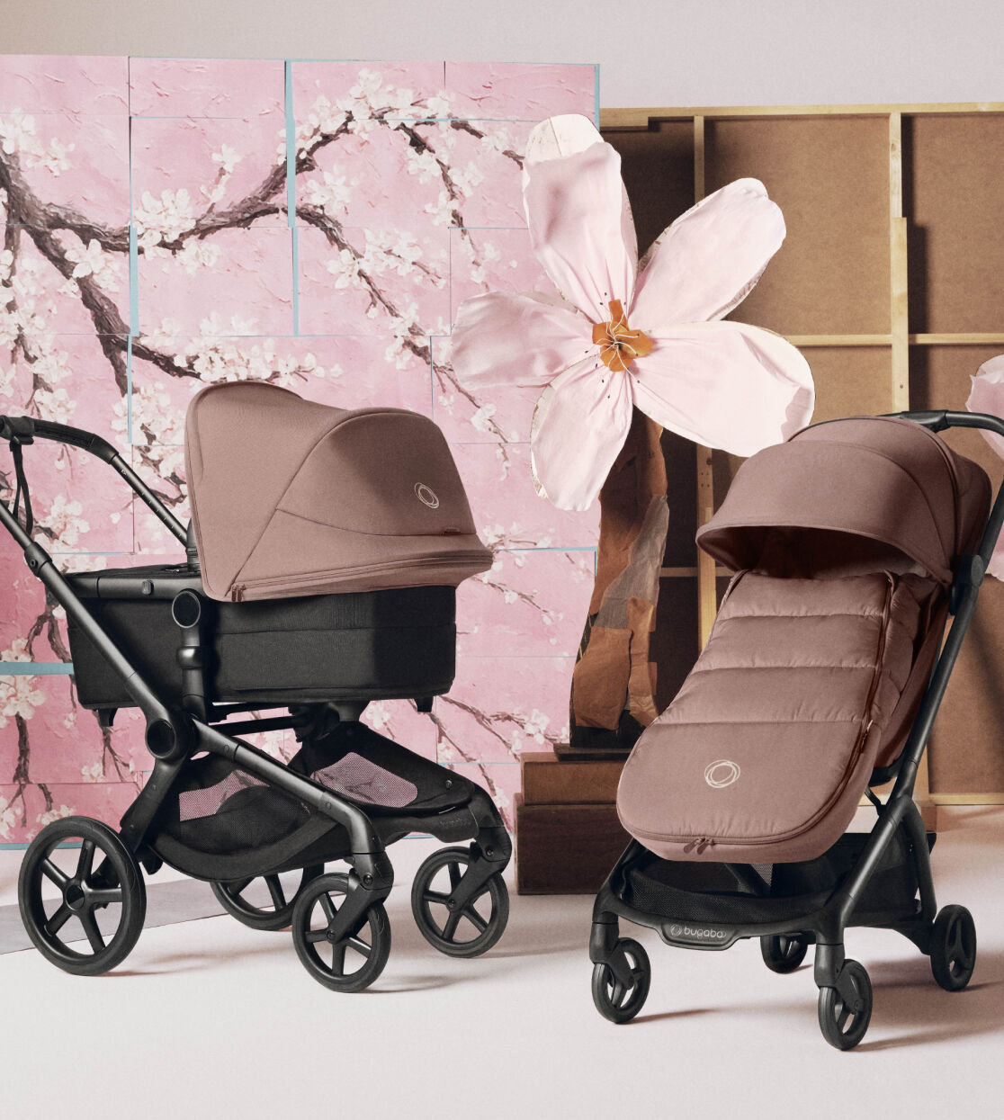 A studio scene with two Bugaboo strollers with Dusty Pink fabrics in the center. On the side, a Bugaboo organizer is hanging on a wooden ladder. They’re displayed in front of a soft pink cherry blossom backdrop and large floral props.