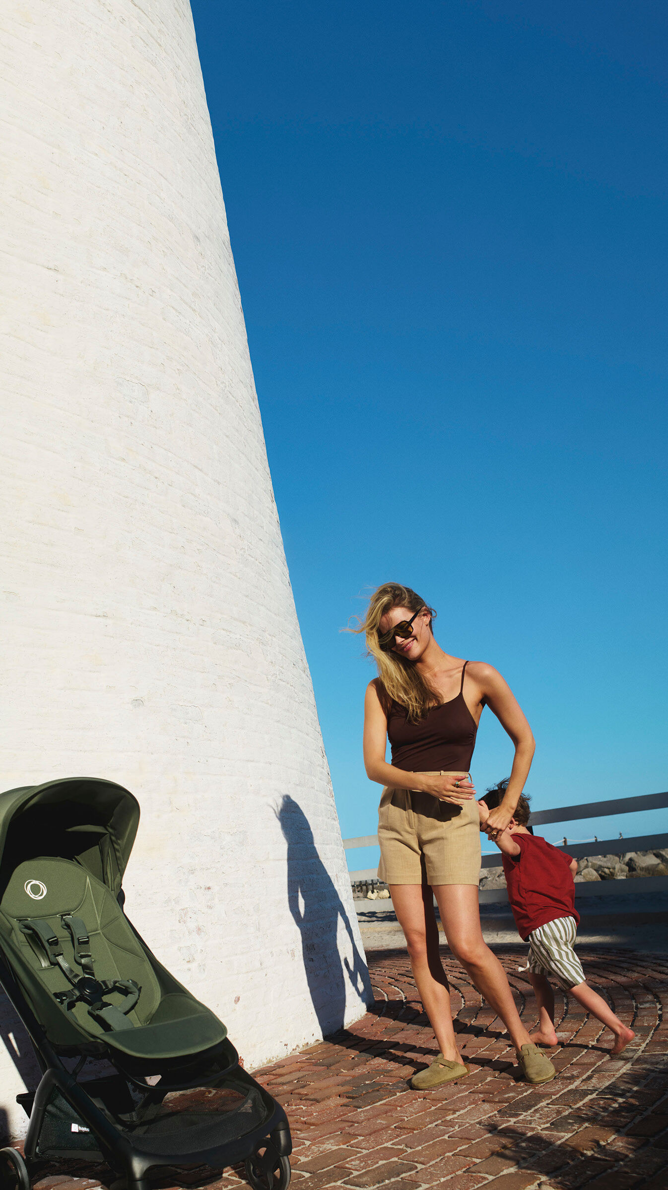 Mom and son walk toward a Bugaboo Butterfly 2 travel stroller in green fabrics next to a white stone building, with a clear blue sky in the background.