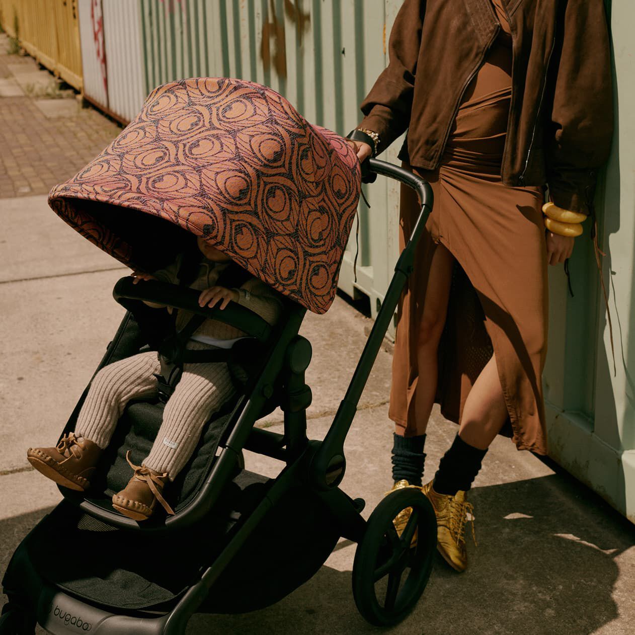 A stylish mom stands outdoors, leaning casually against the side of a metal container. Next to her, her child sits inside a Bugaboo Fox 5 Renew stroller with a Bugaboo X Artipoppe Limited Edition sun canopy.