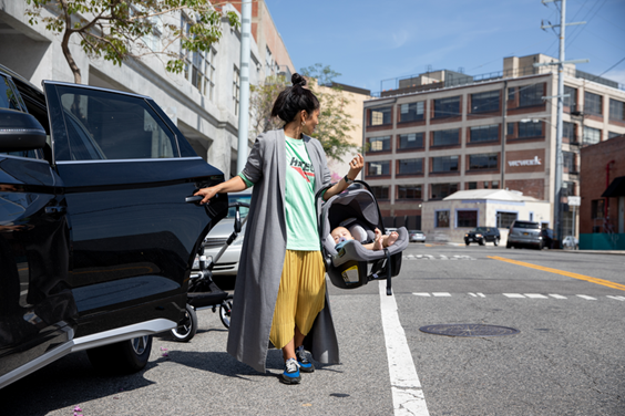 A woman wearing a blue top, yellow trousers and a grey coat carrying a baby sitting in Bugaboo’s Turtle Air by Nuna car seat from the car