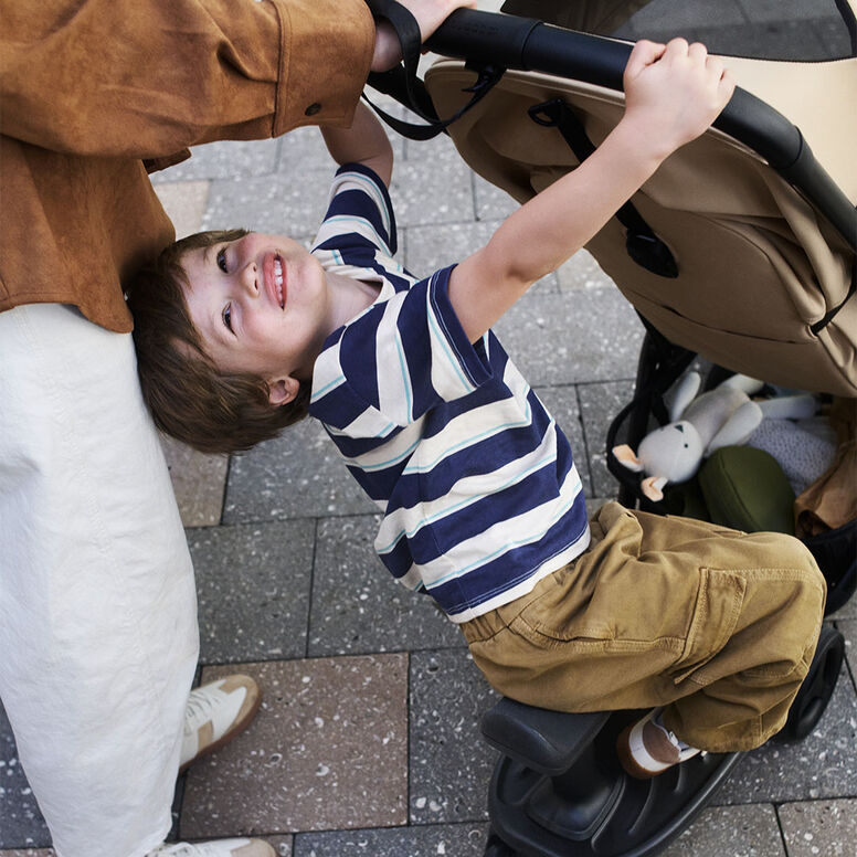 A grinning boy rides on a Bugaboo wheeled board as he clings on to the stroller's handlebar.